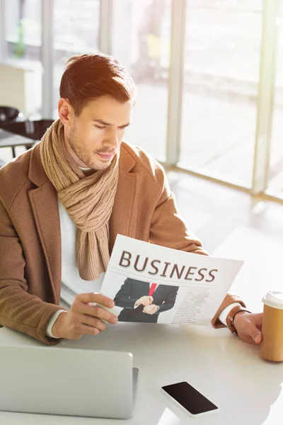 Serious businessman sitting at table, holding paper cup and reading business newspaper in cafe — Stock Photo