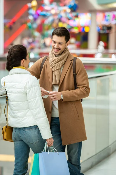 Chica con bolsas de compras hablando con el hombre sonriente en el centro comercial - foto de stock