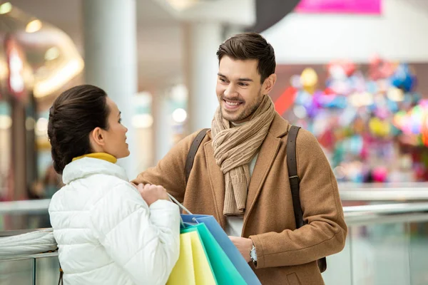 Chica con bolsas de compras hablando con el hombre sonriente en el centro comercial - foto de stock