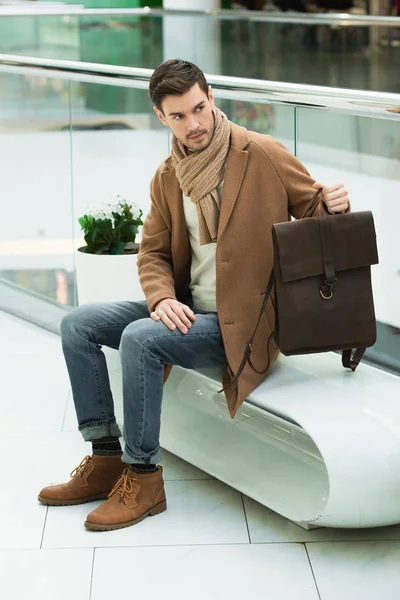 Handsome man holding bag and sitting on bench in shopping mall — Stock Photo