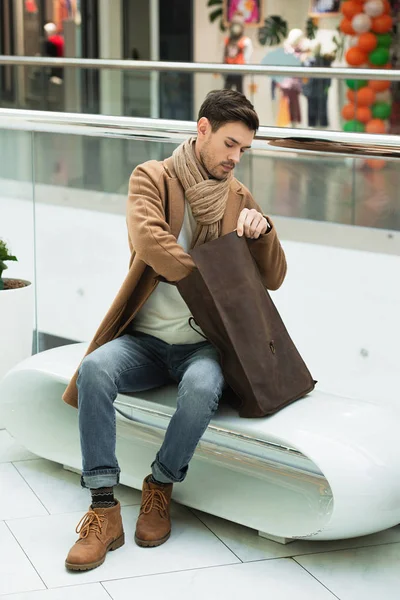 Handsome man sitting on bench and holding bag in shopping mall — Stock Photo