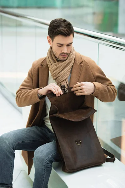 Hombre guapo sentado en el banco y bolsa de apertura en el centro comercial - foto de stock