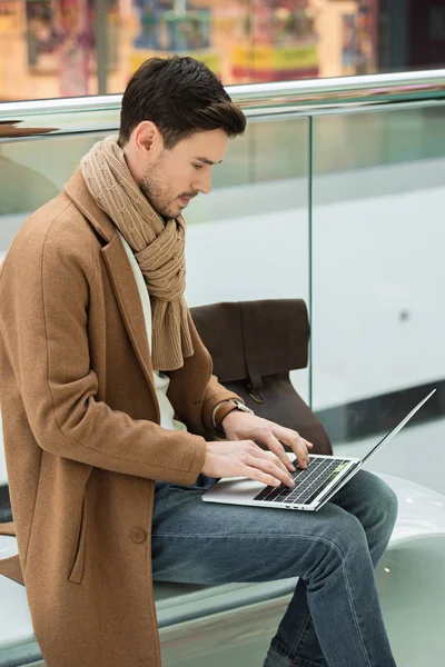 Homem bonito sentado no banco e digitando no teclado do laptop — Fotografia de Stock
