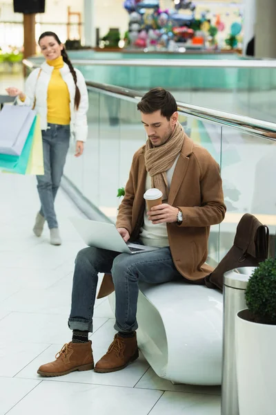 Handsome man sitting on bench,holding disposable cup and typing laptop keyboard in shopping mall — Stock Photo