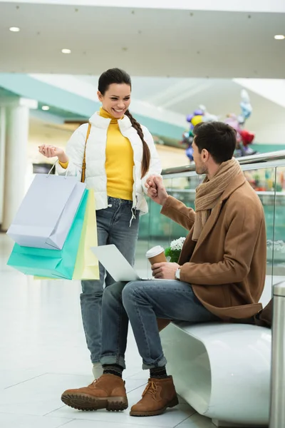 Bel homme avec ordinateur portable et tasse jetable assis sur le banc et tenant la main de la fille avec fille avec des sacs à provisions dans le centre commercial — Photo de stock