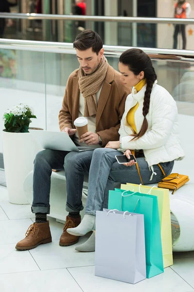 Handsome man with paper cup and girl with smartphone sitting on bench and looking at laptop screen in shopping mall — Stock Photo