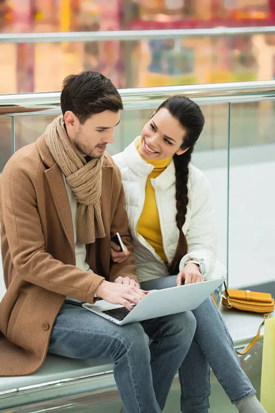 Fille souriante assise sur le banc et regardant bel homme tapant sur le clavier de l'ordinateur portable — Photo de stock