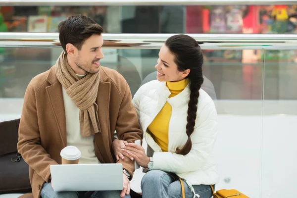 Bel homme avec tasse jetable et ordinateur portable assis sur le banc et regardant fille souriante dans le centre commercial — Photo de stock