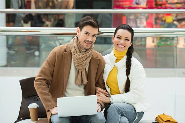 Handsome man with laptop and beautiful girl sitting on bench and looking at camera — Stock Photo