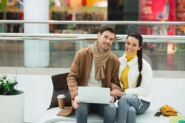 Bel homme avec ordinateur portable et belle fille assise sur le banc et regardant la caméra dans le centre commercial — Photo de stock