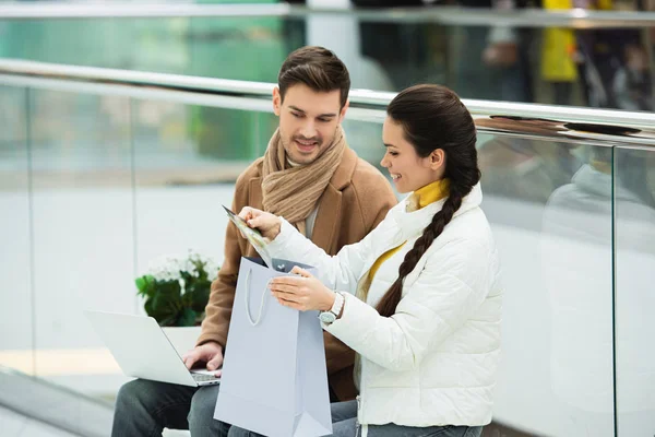 Усміхнений чоловік з ноутбуком і дівчина з сумкою, сидячи на лавці і дивлячись на покупки в торговому центрі — стокове фото