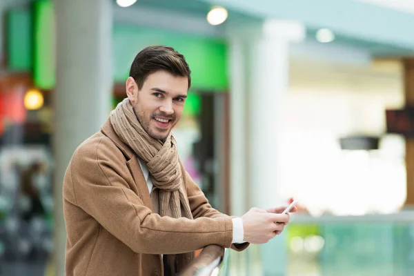 Lächelnder Mann in warmer Kleidung hält Smartphone in der Hand und blickt in die Kamera — Stockfoto