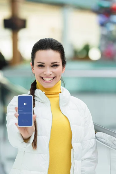 Chica atractiva sonriendo y mostrando la pantalla del teléfono inteligente con aparato de facebook - foto de stock