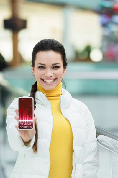 Bella ragazza sorridente, guardando la fotocamera e mostrando lo schermo dello smartphone con l'app corsi di trading — Foto stock