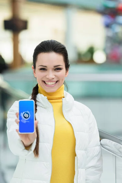 Schöne Mädchen lächelt, blickt in die Kamera und zeigt Smartphone-Bildschirm mit Shazam-App — Stockfoto