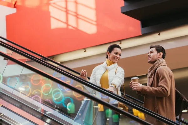 Sorridente ragazza con borse della spesa e bello uomo con tazza usa e getta salendo sulla scala mobile nel centro commerciale — Foto stock