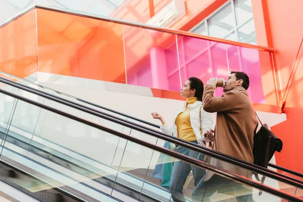 Handsome man drinking coffee from disposable cup and attractive girl with shopping bags on escalator in mall — Stock Photo