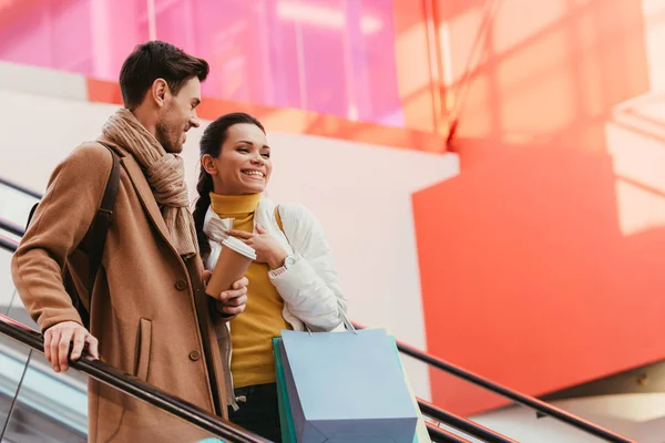 Attraktives Mädchen mit Einkaufstaschen und Mann mit Einwegbecher auf Rolltreppe — Stockfoto
