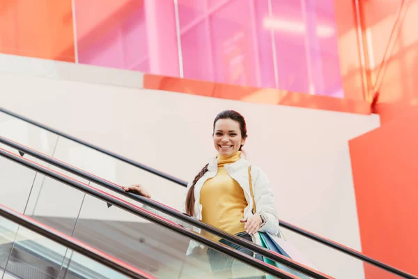 Attractive girl with shopping bags smiling and looking at camera on escalator — Stock Photo