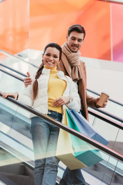 Hombre guapo con taza de papel abrazando chica atractiva con bolsas de compras en escaleras mecánicas - foto de stock