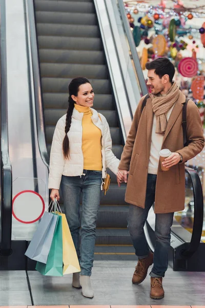 Hombre guapo con taza de papel y chica atractiva con bolsas de compras cogidas de la mano y mirando unos a otros cerca de escaleras mecánicas - foto de stock