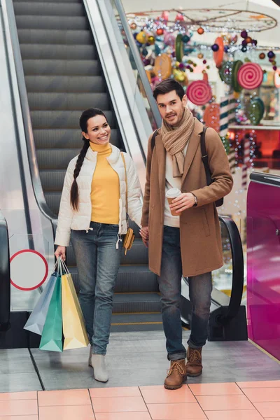 Chica atractiva con bolsas de compras de la mano con el hombre guapo mirando a la cámara cerca de escaleras mecánicas - foto de stock