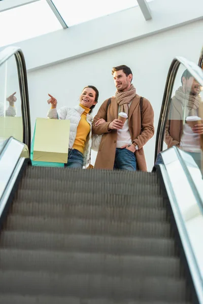 Chica atractiva con bolsas de compras apuntando con el dedo y la mano de hombre guapo con taza de papel en la parte superior de escaleras mecánicas - foto de stock