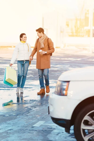 Couple aimant avec des sacs à provisions et une tasse jetable tenant la main et passant par le parking — Photo de stock