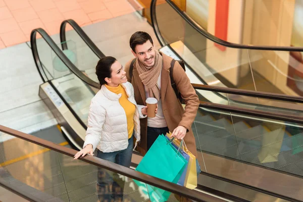 Hombre sonriente sosteniendo bolsas de compras y chica atractiva con taza desechable en escaleras mecánicas - foto de stock
