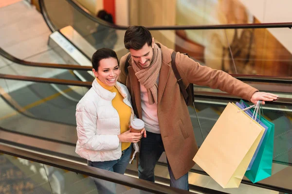 Bel homme avec des sacs à provisions et jolie fille avec tasse jetable monter sur escalator — Photo de stock