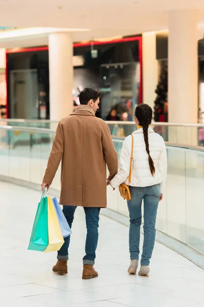 Vista trasera de pareja amorosa con bolsas de compras cogidas de la mano en el centro comercial - foto de stock