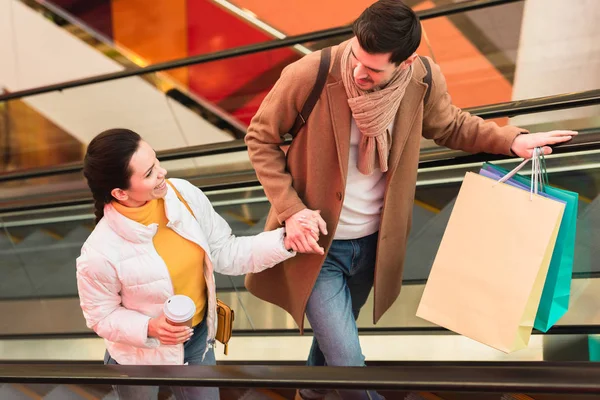 Hombre sonriente con bolsas de compras y hermosa chica con taza desechable mirándose el uno al otro en escaleras mecánicas - foto de stock