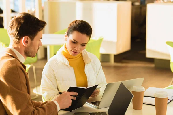 Hombre guapo mostrando cuaderno a chica atractiva en la mesa en la cafetería - foto de stock