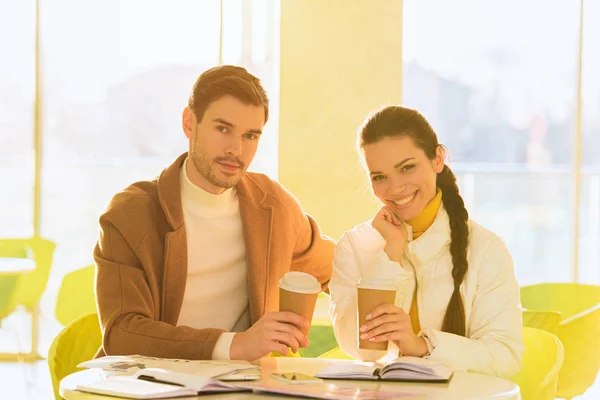 Bel homme et fille souriante tenant des tasses jetables et regardant la caméra dans le café — Photo de stock