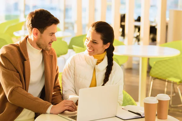 Handsome man and smiling girl sitting at table with laptop and looking at each other in cafe — Stock Photo