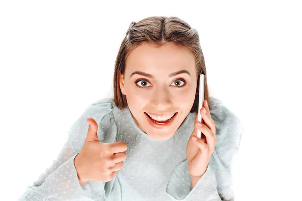 Vista de ángulo alto de la mujer sonriente que muestra el pulgar hacia arriba mientras habla en el teléfono inteligente aislado en blanco - foto de stock