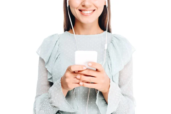 Recortado disparo de la mujer sonriente usando teléfono inteligente mientras escucha música en auriculares aislados en blanco - foto de stock