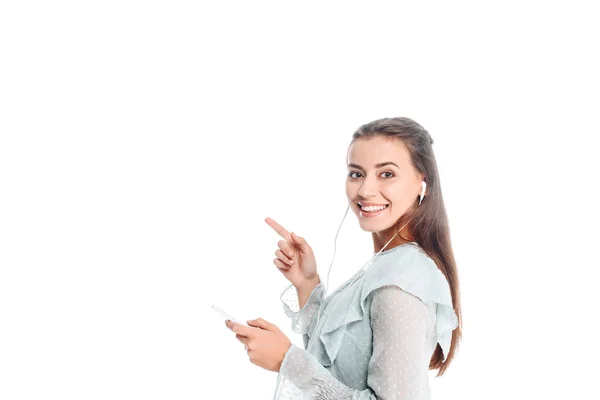 Vue latérale de la femme souriante avec smartphone pointant du doigt tout en écoutant de la musique dans des écouteurs isolés sur blanc — Photo de stock