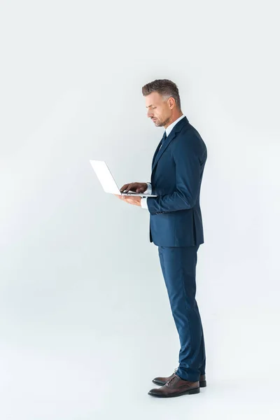 Vista lateral de empresário bonito em terno usando laptop isolado no branco — Fotografia de Stock