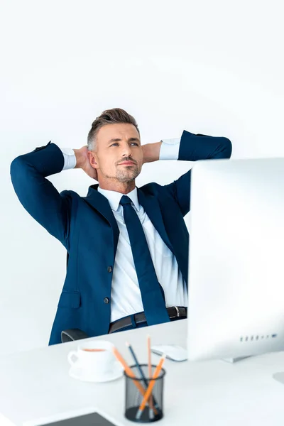 Handsome businessman in suit sitting at table with computer and holding hands behind head isolated on white — Stock Photo