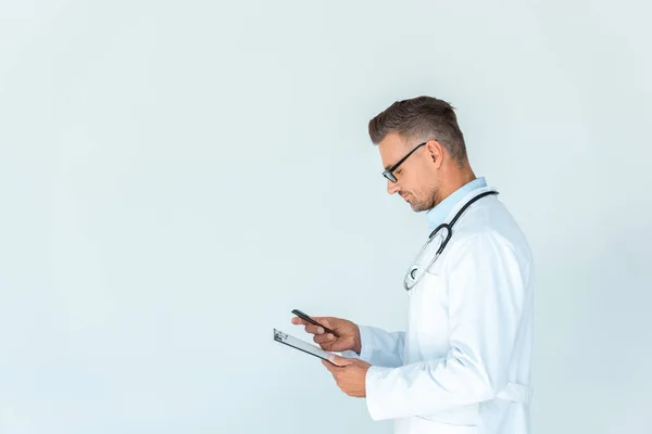 Vue latérale du beau médecin dans des lunettes avec stéthoscope sur les épaules tenant presse-papiers et smartphone isolé sur blanc — Photo de stock