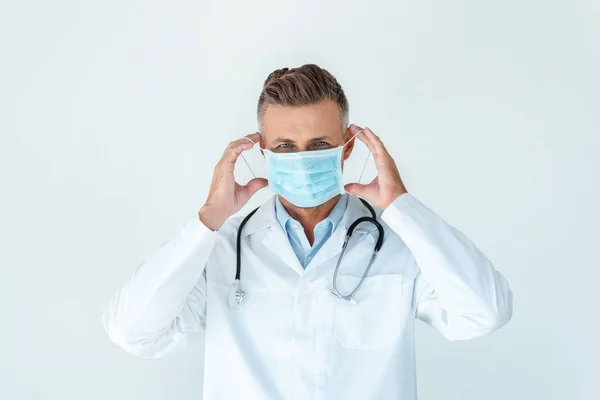 Красивый врач в медицинской маске и глядя на камеру, изолированную на белом — стоковое фото