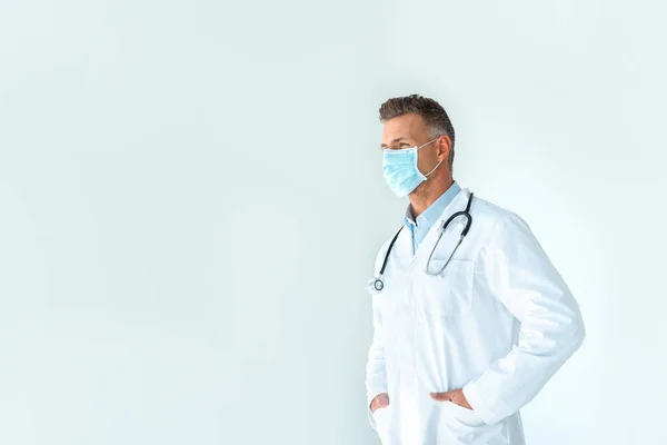 Guapo doctor en bata blanca y máscara médica mirando hacia otro lado aislado en blanco - foto de stock