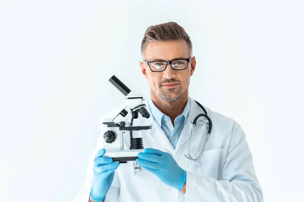 Handsome scientist in glasses holding microscope and looking at camera isolated on white — Stock Photo