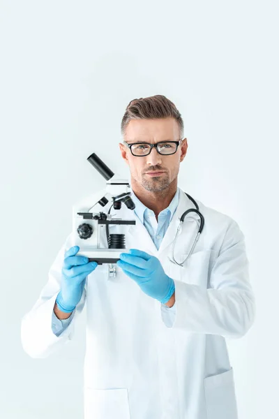 Handsome serious scientist in glasses holding microscope and looking at camera isolated on white — Stock Photo