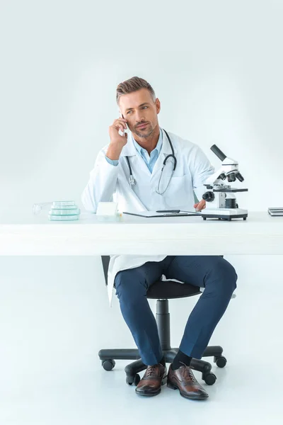 Beau scientifique parlant par smartphone à table avec microscope isolé sur blanc — Photo de stock