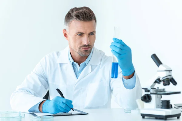 Красивий вчений дивиться на пробірку з синім реагентом ізольовано на білому — стокове фото