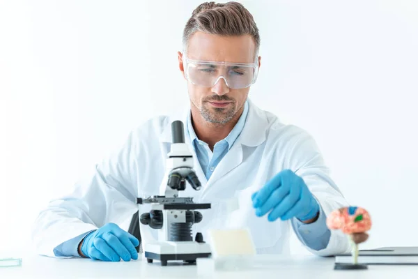 Foco seletivo do cientista bonito em óculos de proteção fazendo experiência com microscópio isolado em branco — Fotografia de Stock