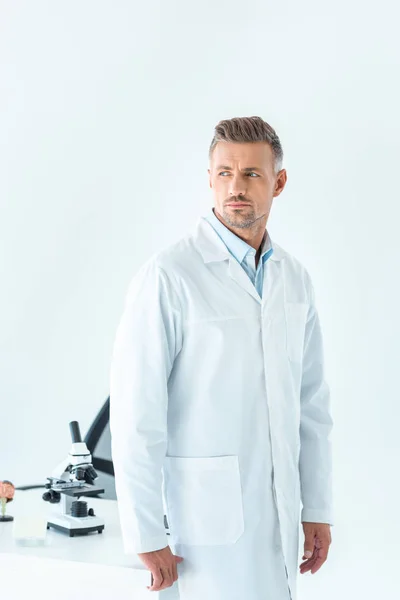 Красивый ученый, стоящий рядом с столом с микроскопом и глядя в сторону изолированы на белом — стоковое фото