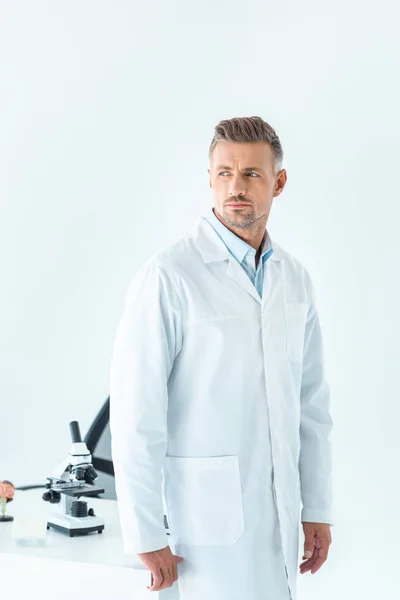 Handsome scientist standing near table with microscope and looking away isolated on white — Stock Photo